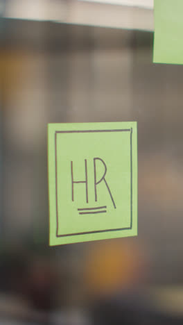 Vertical-Video-Close-Up-Of-Woman-Putting-Sticky-Note-With-HR-Written-On-It-Onto-Transparent-Screen-In-Office-